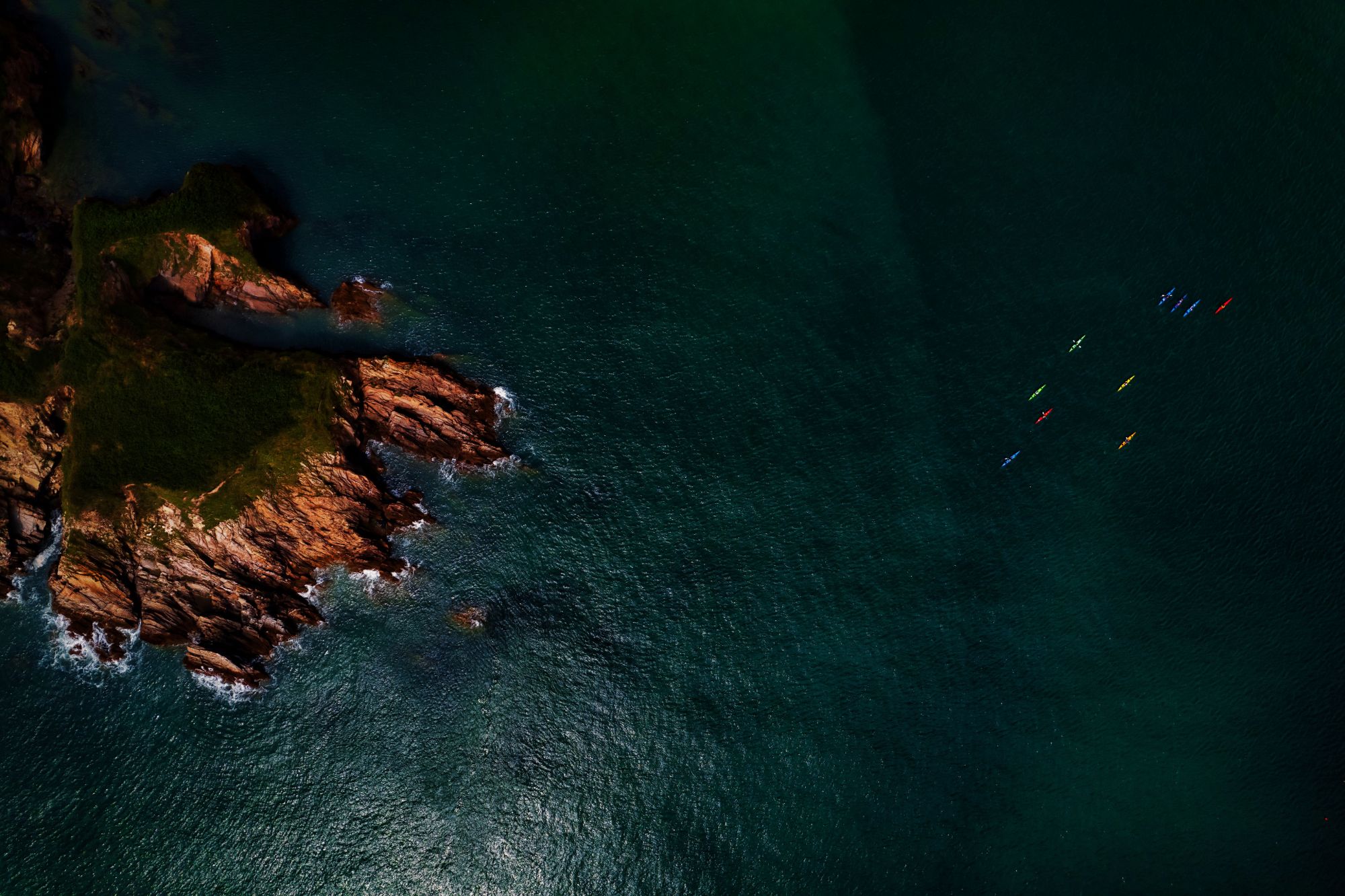 Aerial view of a rocky green headland meeting deep teal water, with a group of kayaks offshore